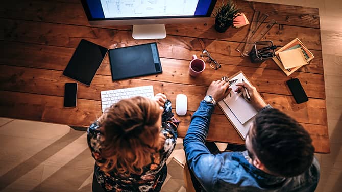 Os significados de violação de dados e vazamento de dados podem muitas vezes ser confundidos, mas os dois não são exatamente a mesma coisa. A imagem mostra duas pessoas sentadas em um computador, uma tomando notas.