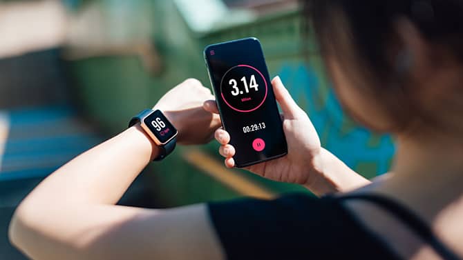 Uma mulher usando um aplicativo de fitness em seu smartphone e relógio inteligente. Os aplicativos de monitoramento de fitness são um exemplo dos potenciais efeitos positivos da tecnologia na saúde.